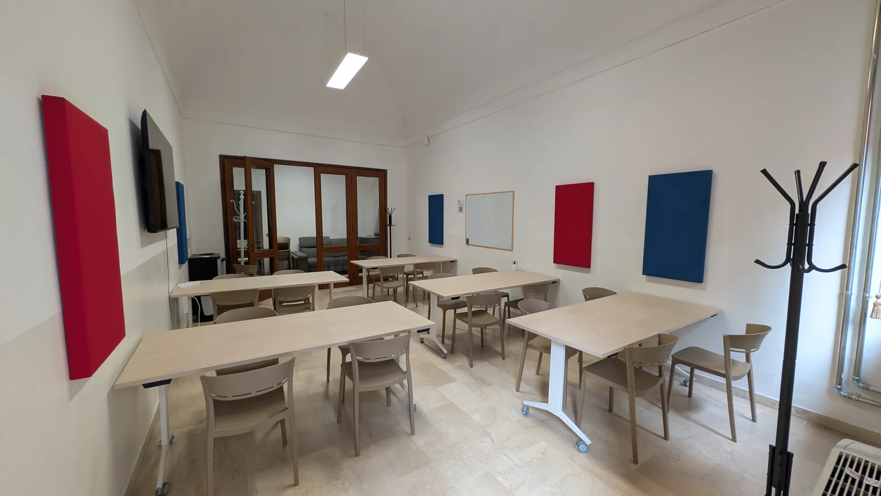 Aula 1: pavimento in marmo, tavoli chiari, pannelli rossi e blu alle pareti e una porta a vetri in legno.