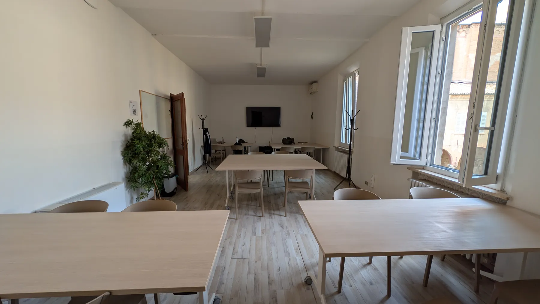 Aula 2: tavoli chiari in fila, parquet, due finestre grandi aperte e una pianta.