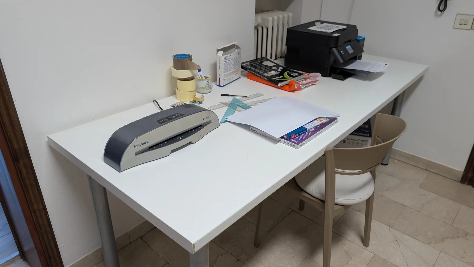 L’angolo cartoleria: una scrivania bianca con stampante, plastificatrice, nastro adesivo e quaderni.