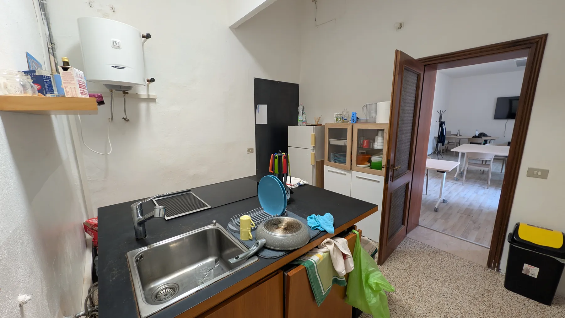 La cucina: lavello in acciaio, piano di lavoro nero, pensili e una porta aperta verso un’aula.
