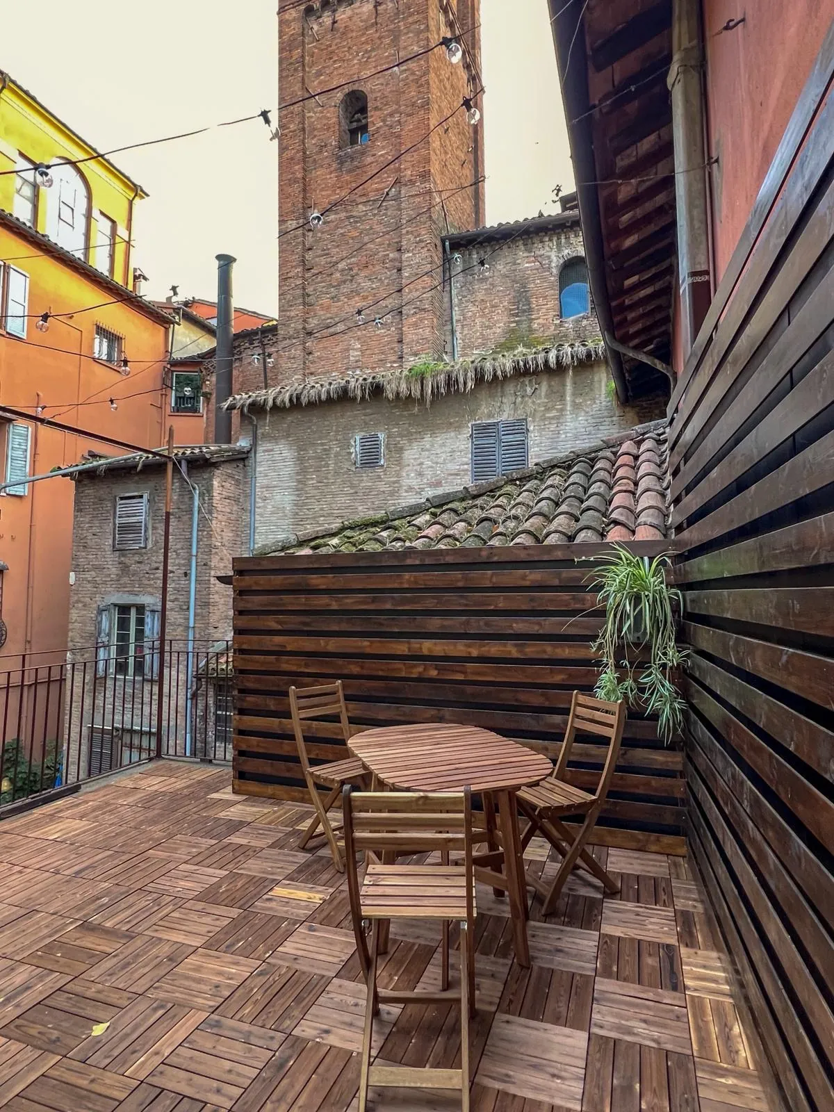 Il terrazzo di giorno: pavimento in legno, un tavolo con quattro sedie, una parete di doghe e il campanile dietro i tetti.